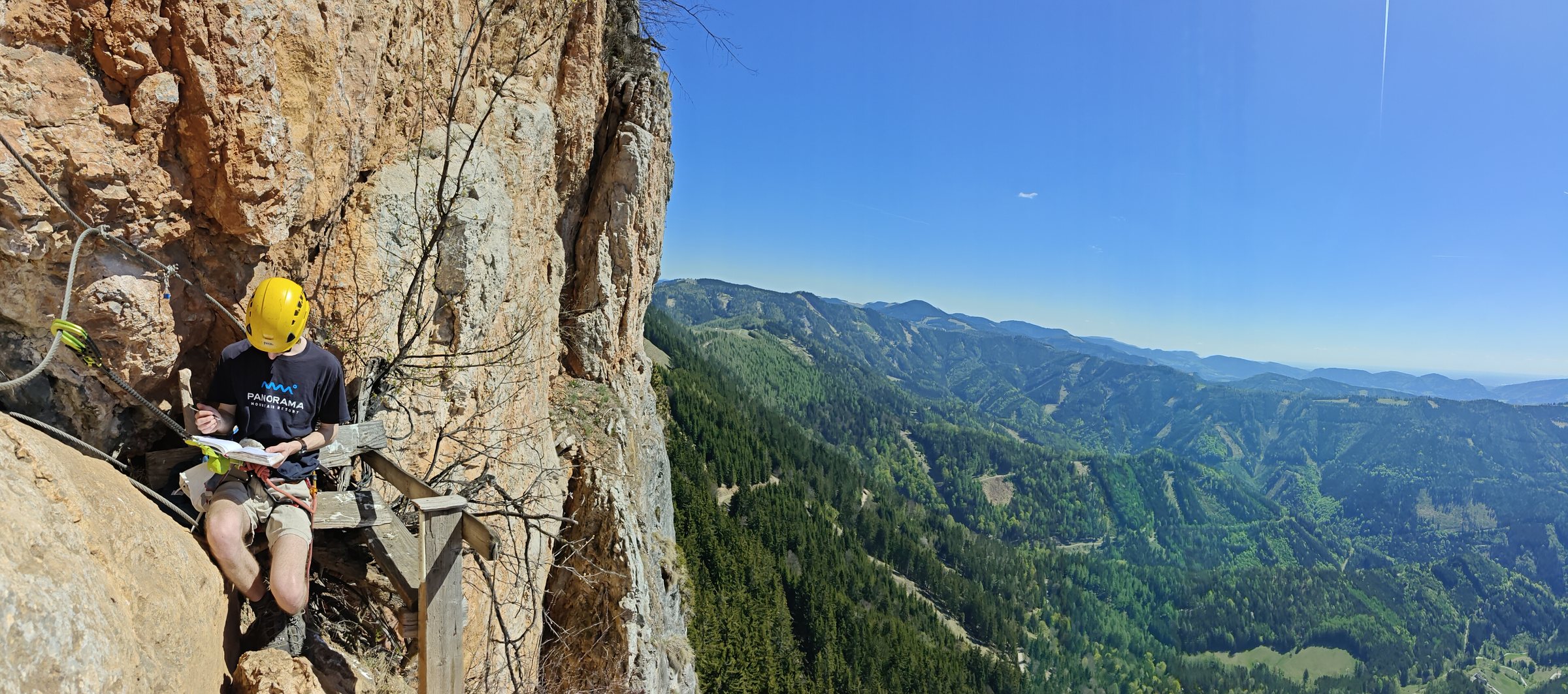 A climber clipped into a via ferrata, sitting on a wooden ledge partway up the face, reading, with the valley below
